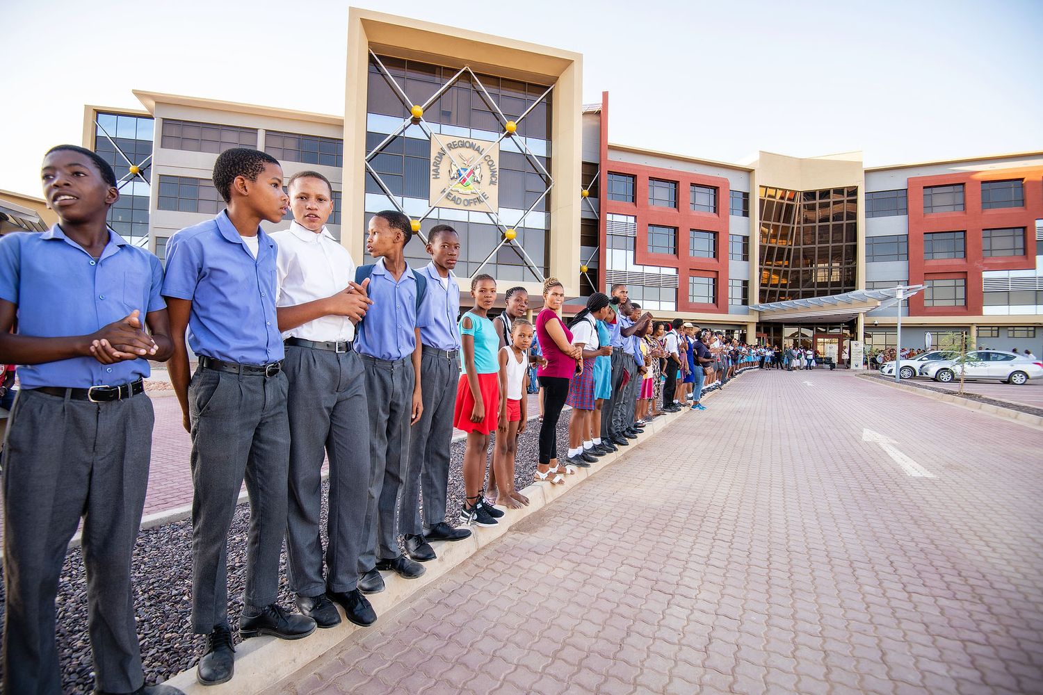 Students in school uniforms standing in a line outside a modern school building on a sunny day.