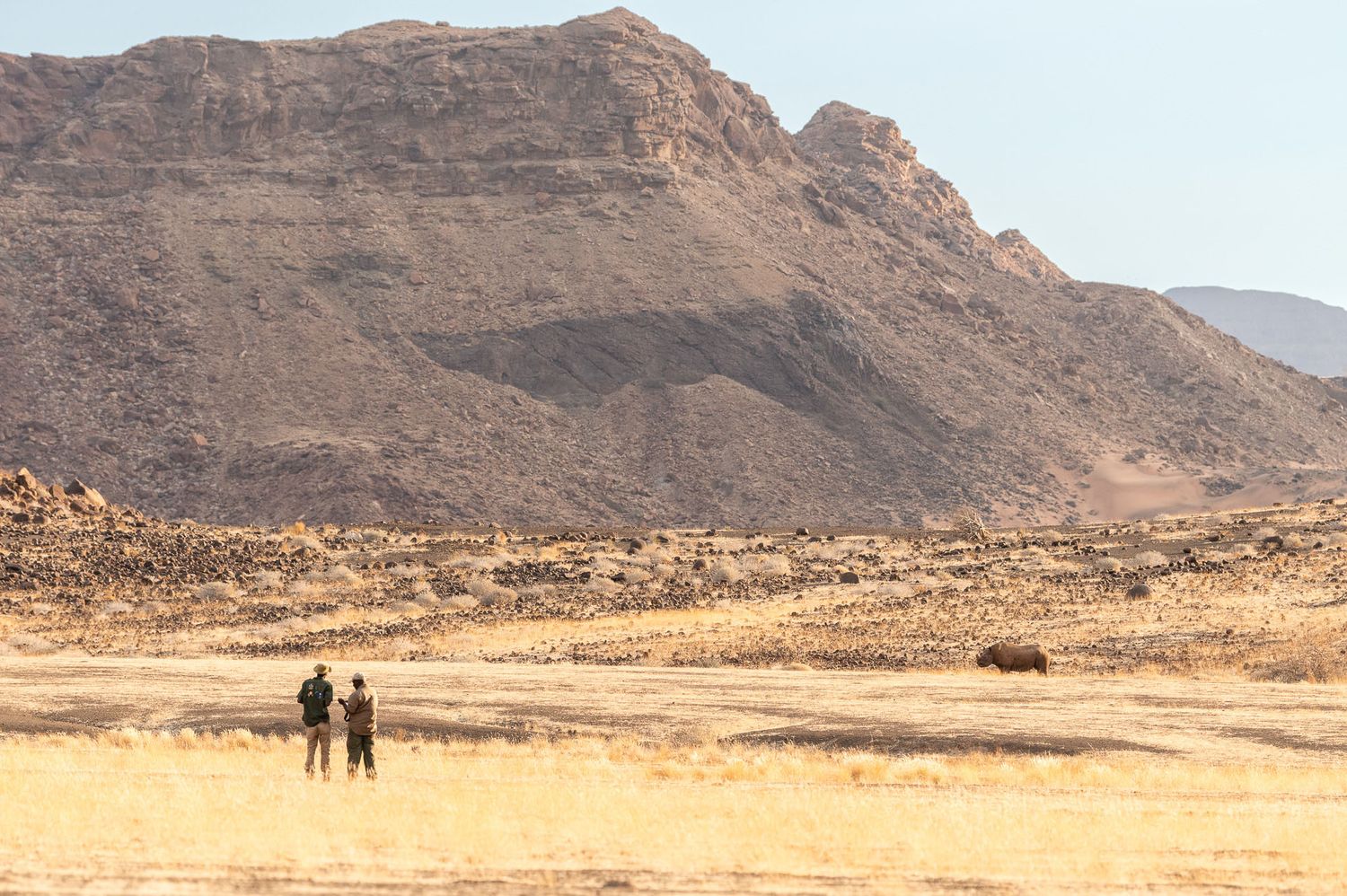 Two people stand in a vast arid desert landscape with a rocky mountain backdrop under clear blue skies.