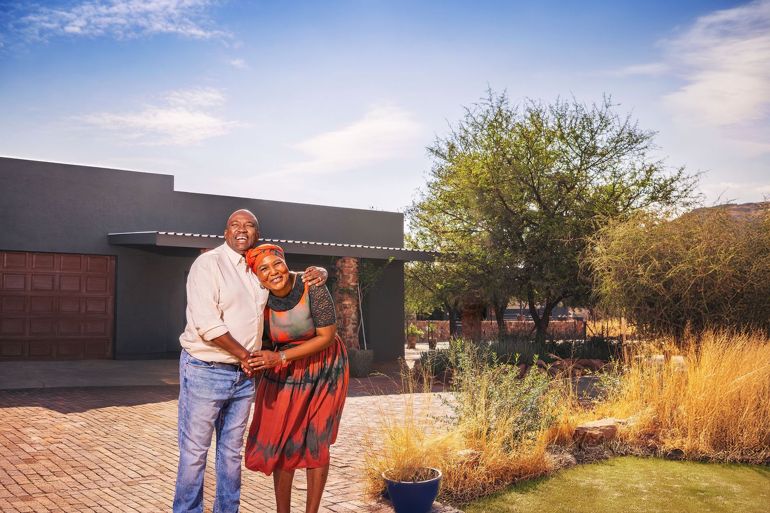 A smiling couple embraces outdoors in front of a modern home surrounded by lush garden greenery under a blue sky.