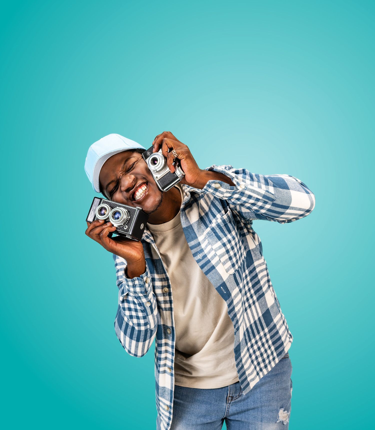 Young man in plaid shirt and white cap holding two vintage cameras, smiling against a teal background.