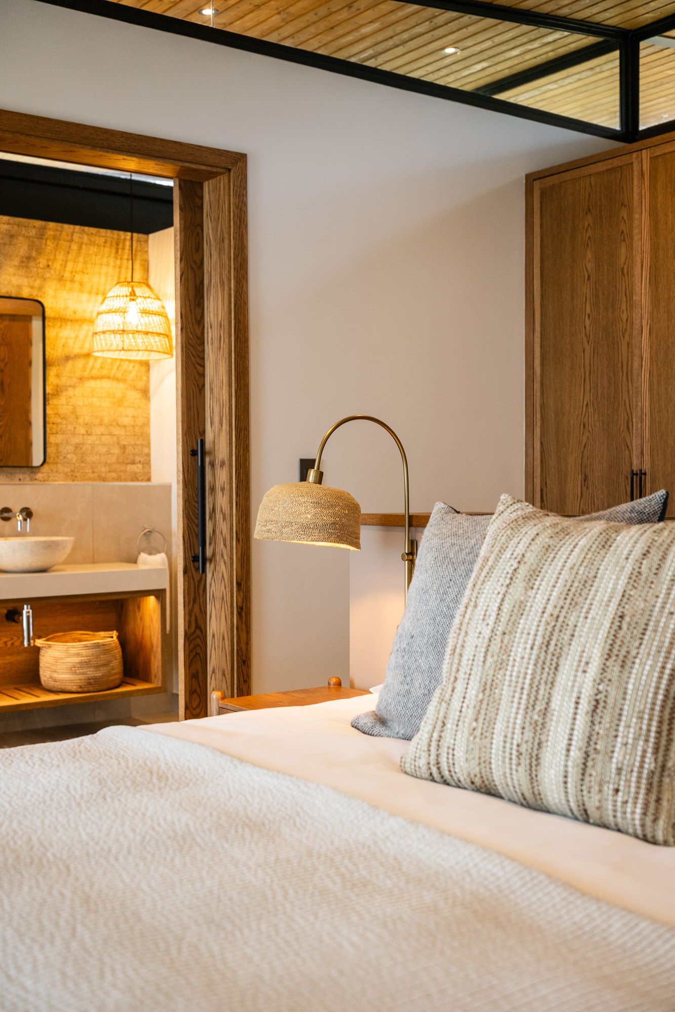 Elegant bedroom with white bedding, woven bedside lamp, and en-suite bathroom with vessel sink visible.