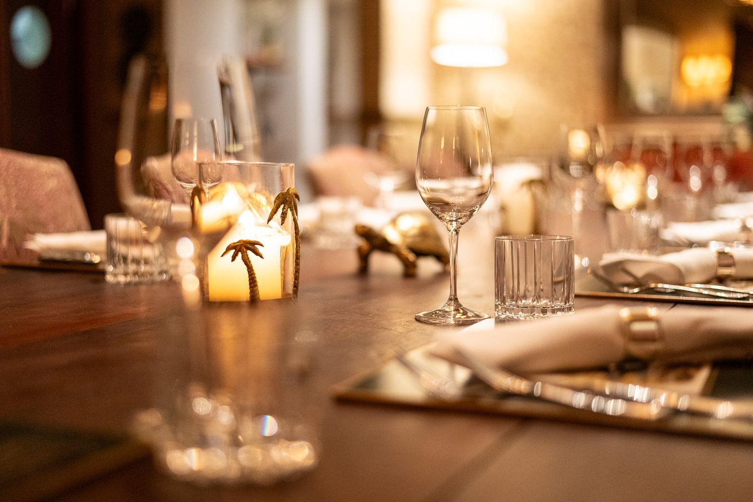 Elegant dining table set with crystal wine glasses, candles, and golden palm tree decor in warm ambient lighting.
