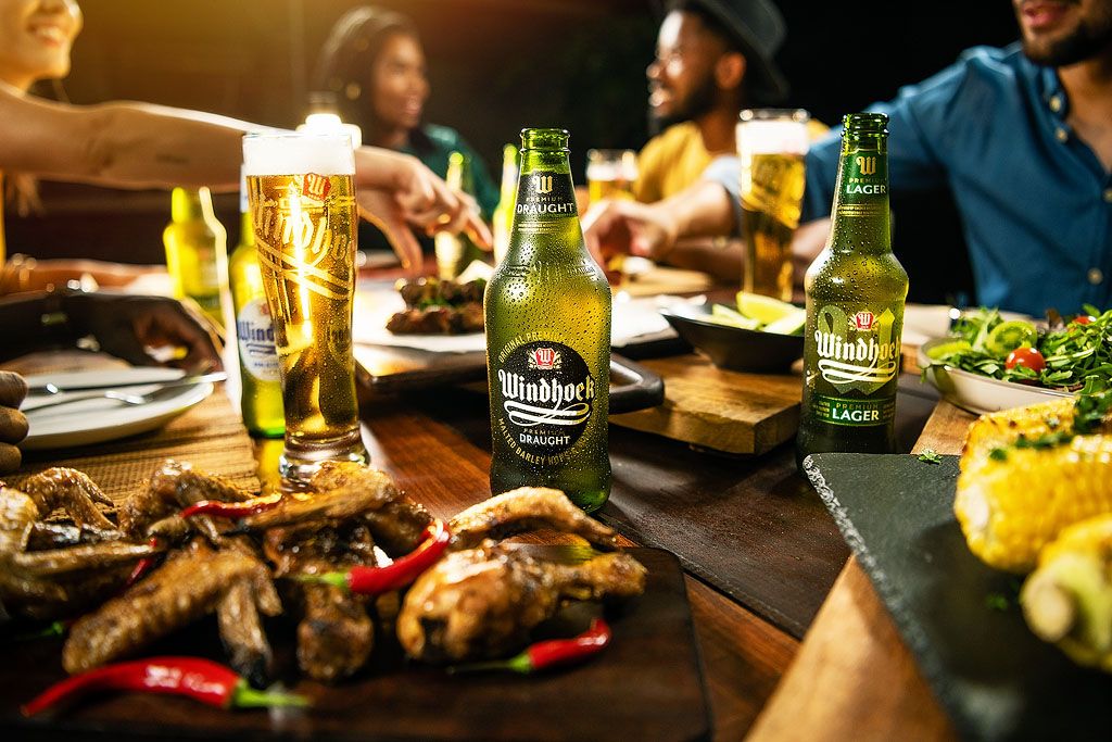 Friends enjoying Windhoek Lager beer with grilled chicken and corn at a restaurant dinner table.