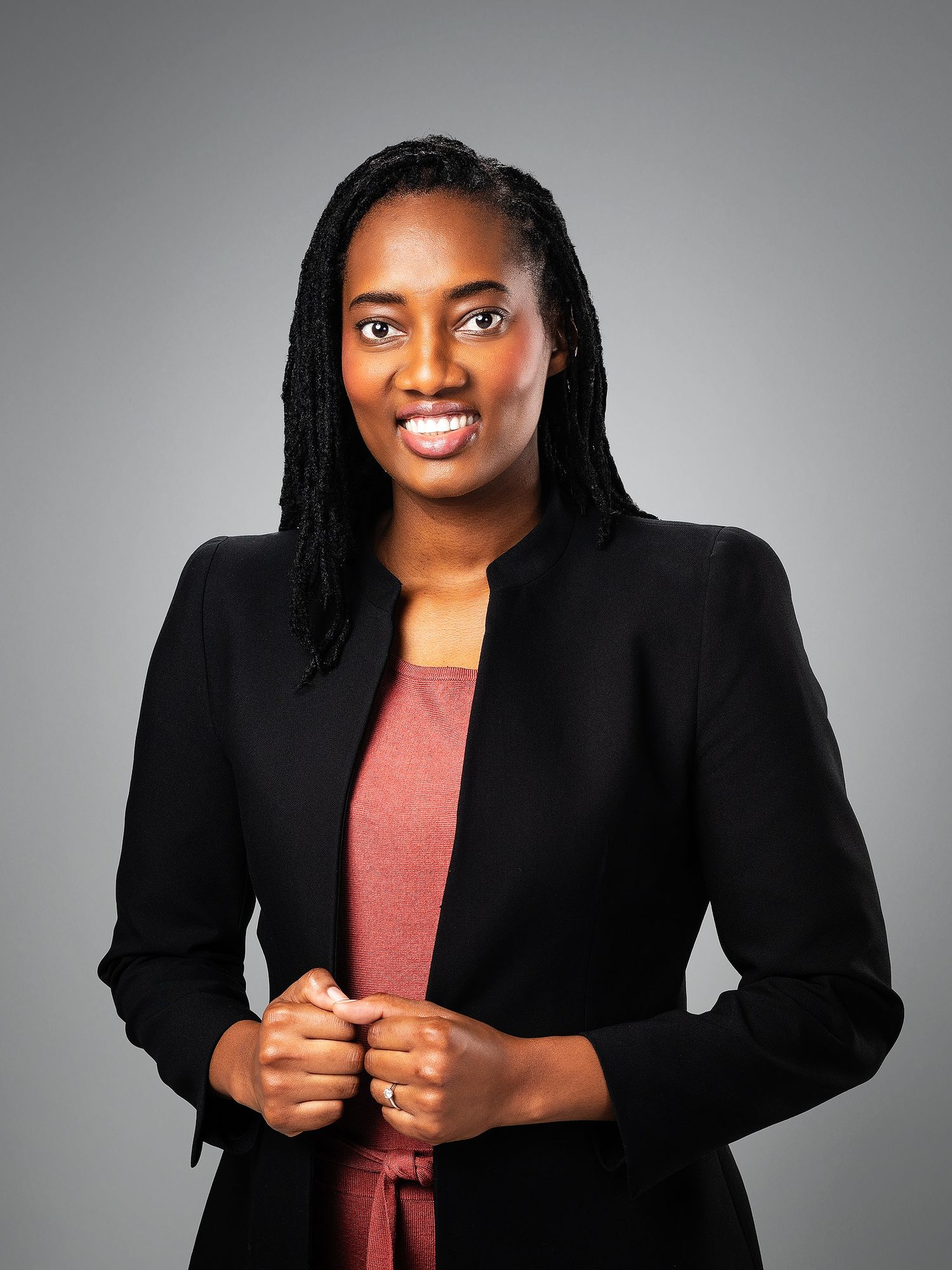Professional woman in black blazer and pink top smiling confidently against gray background.