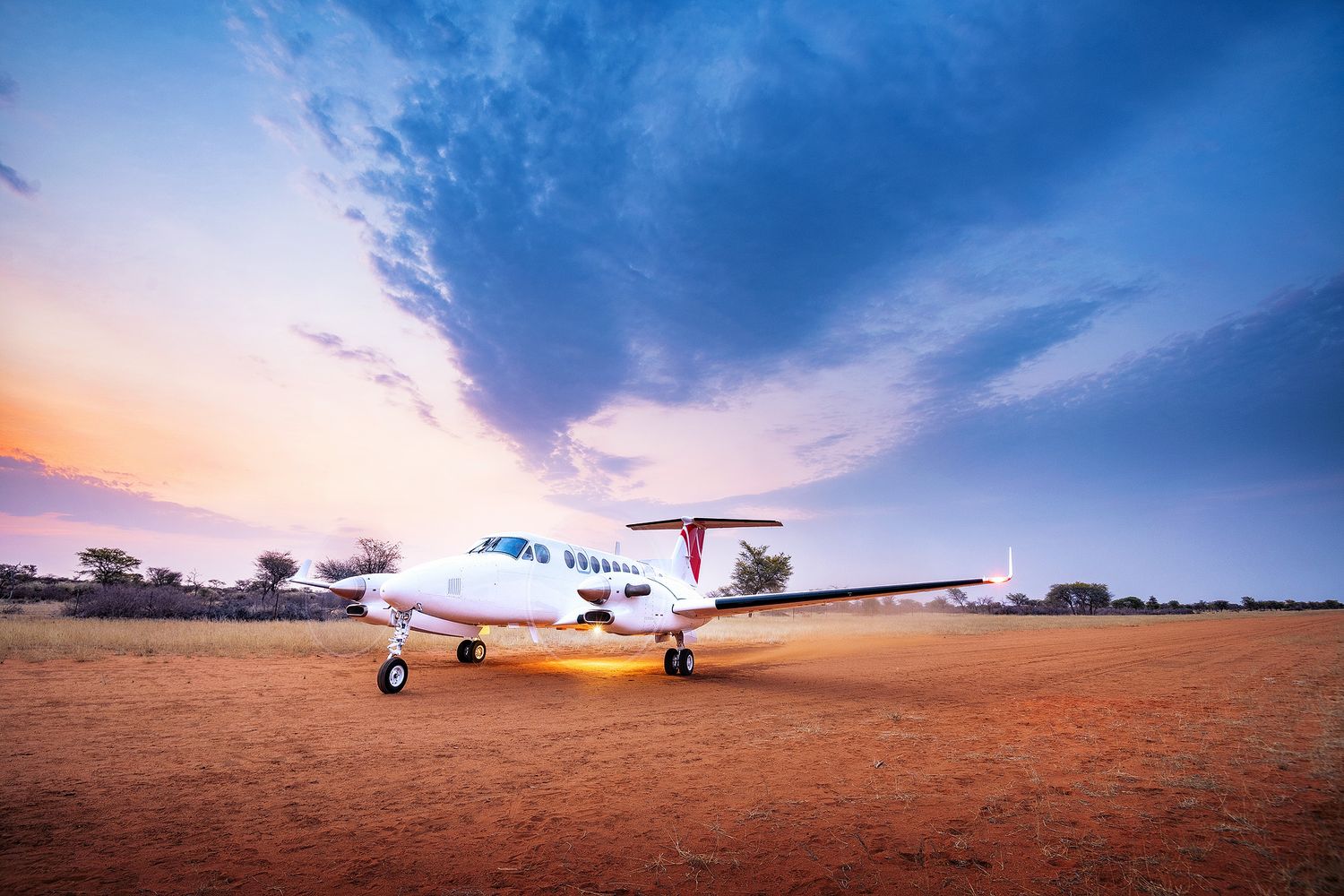 Small propeller plane parked on red dirt airstrip at sunset with dramatic colorful sky in remote Australian outback.
