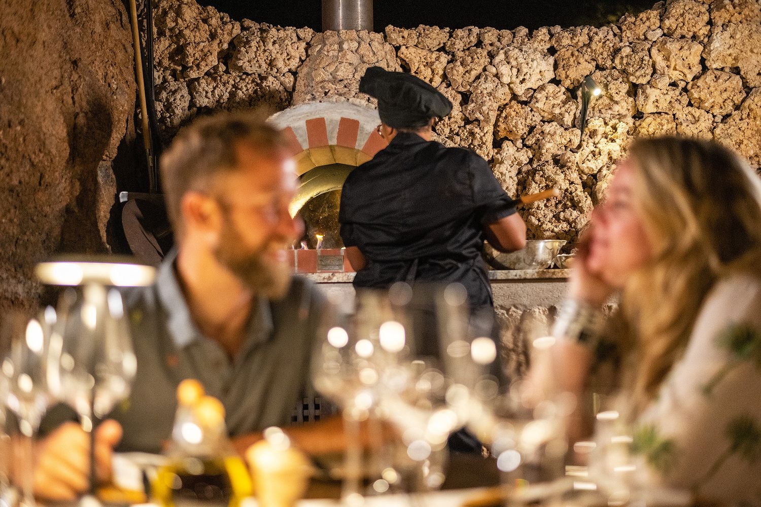 Couple dining at a rustic restaurant with a wood-fired pizza oven and chef in the background.