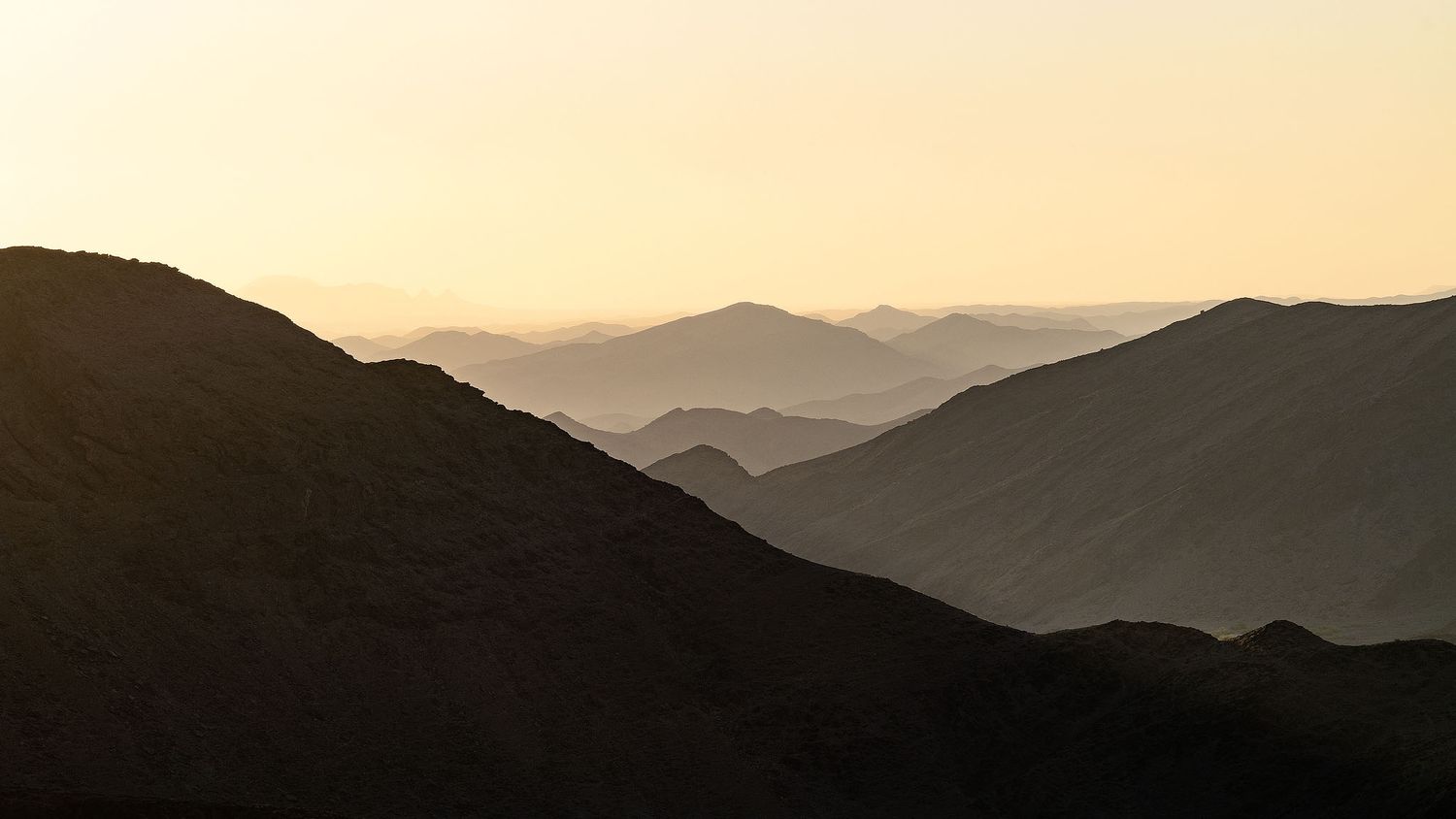Silhouetted mountain ridges layered in hazy golden sunset light, creating a serene and dramatic landscape panorama.