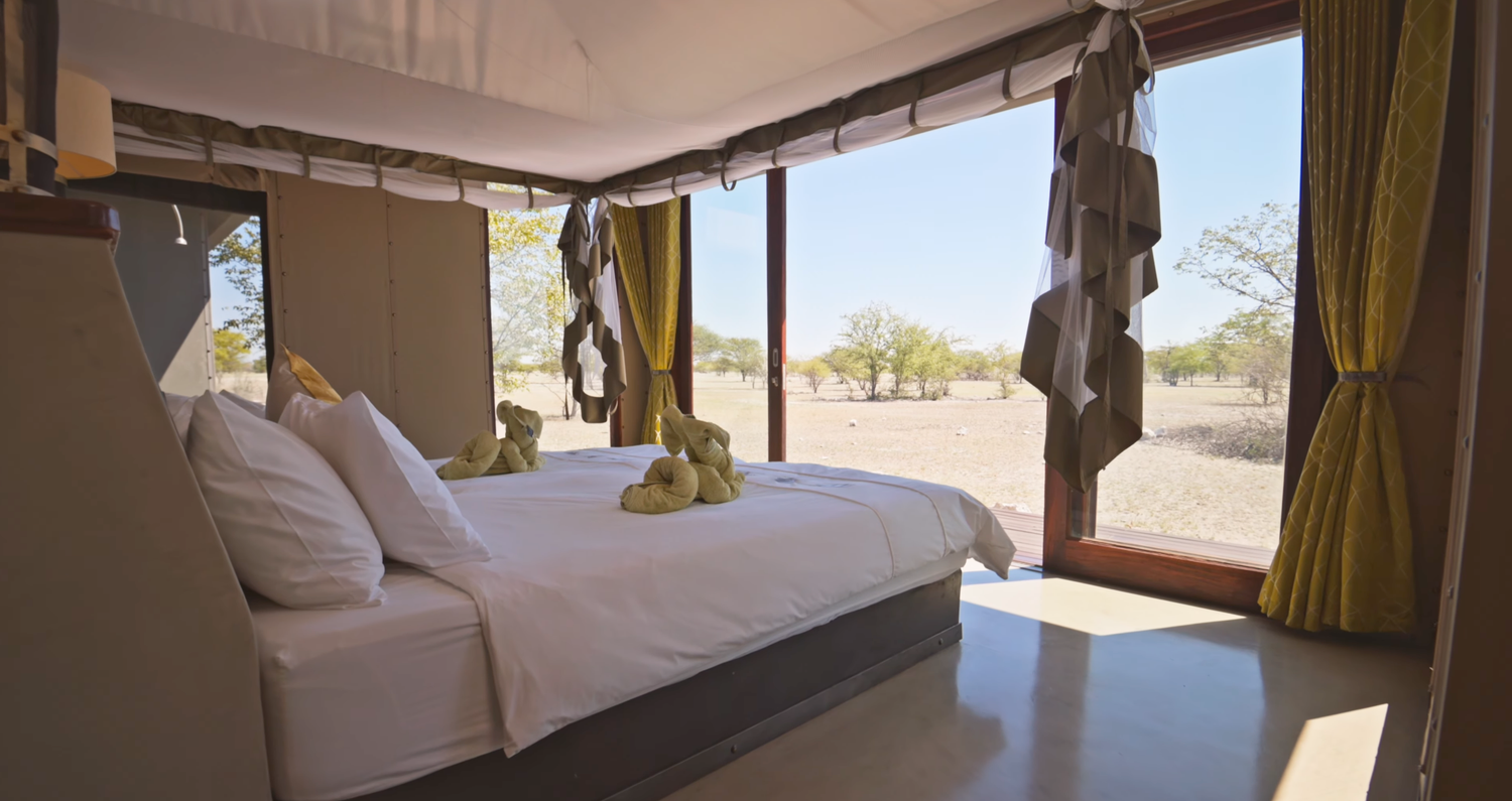 Luxury safari tent bedroom with white bedding, yellow curtains, and panoramic views of African savanna landscape.