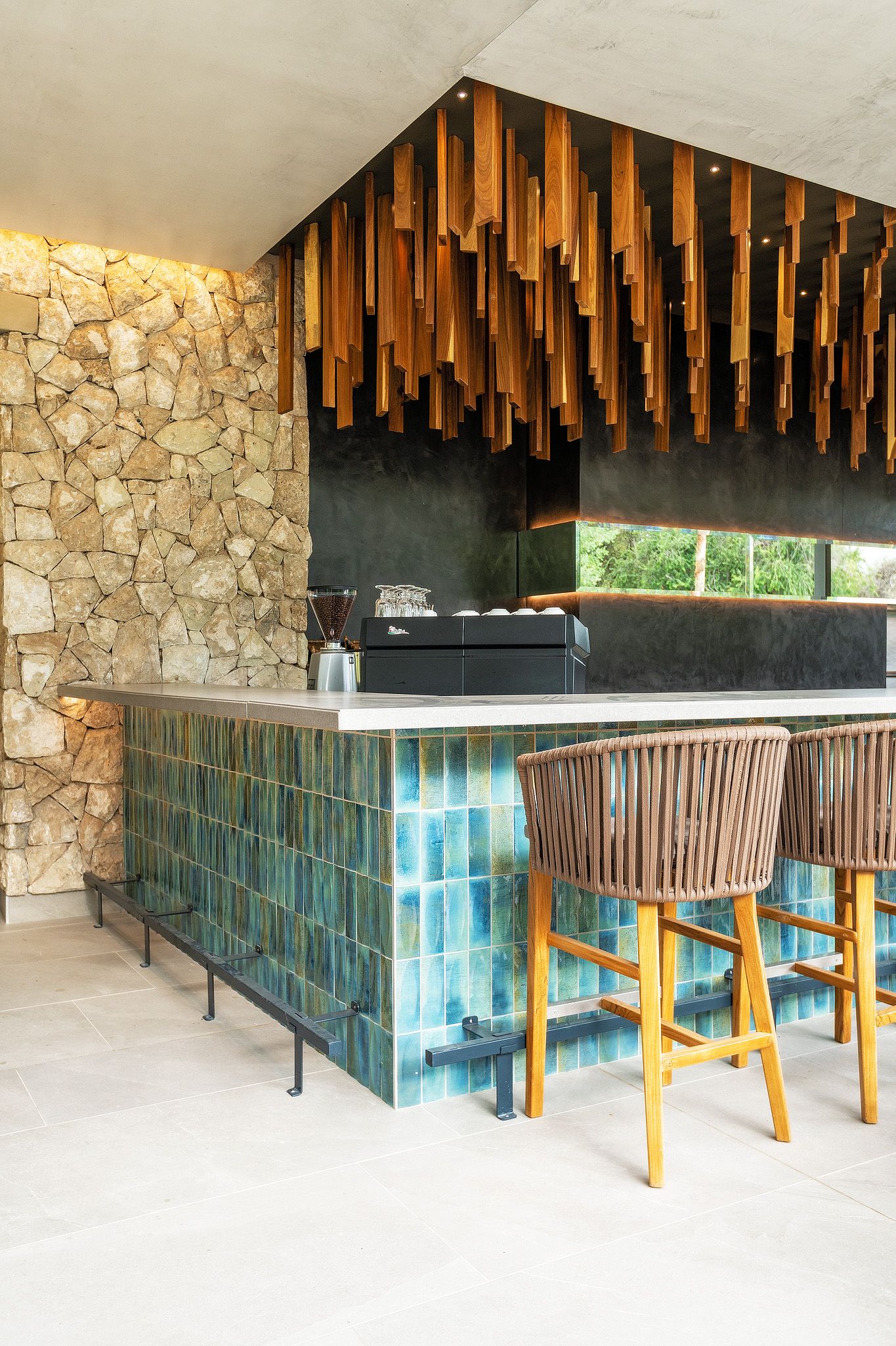 Modern bar with teal tiles, wicker stools, stone wall, and hanging wooden ceiling installation.