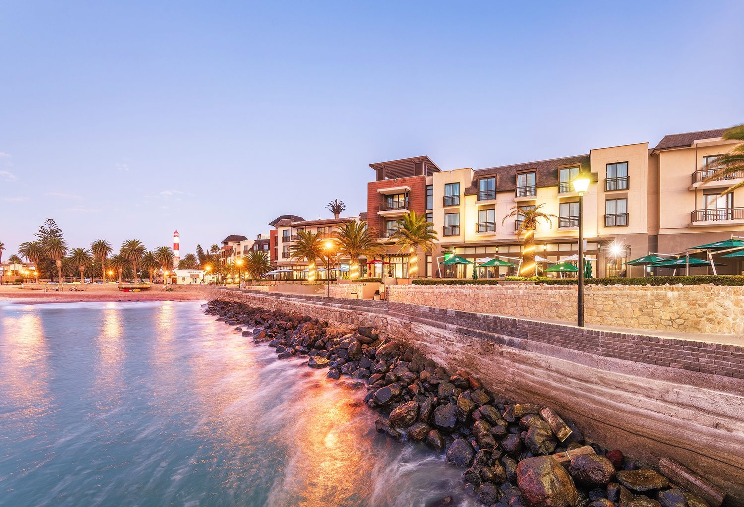 Elegant coastal hotel and seafront promenade glowing at dusk with rocky breakwater and serene blue water reflections.