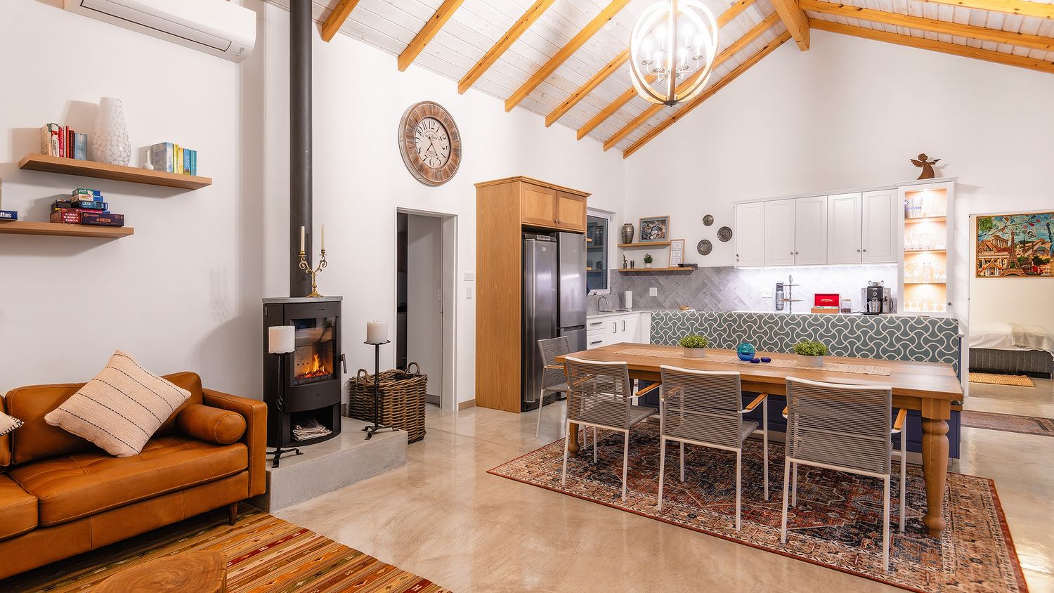 Open-plan living area with wood-burning stove, leather sofa, dining table, and modern kitchen with vaulted wooden ceiling.
