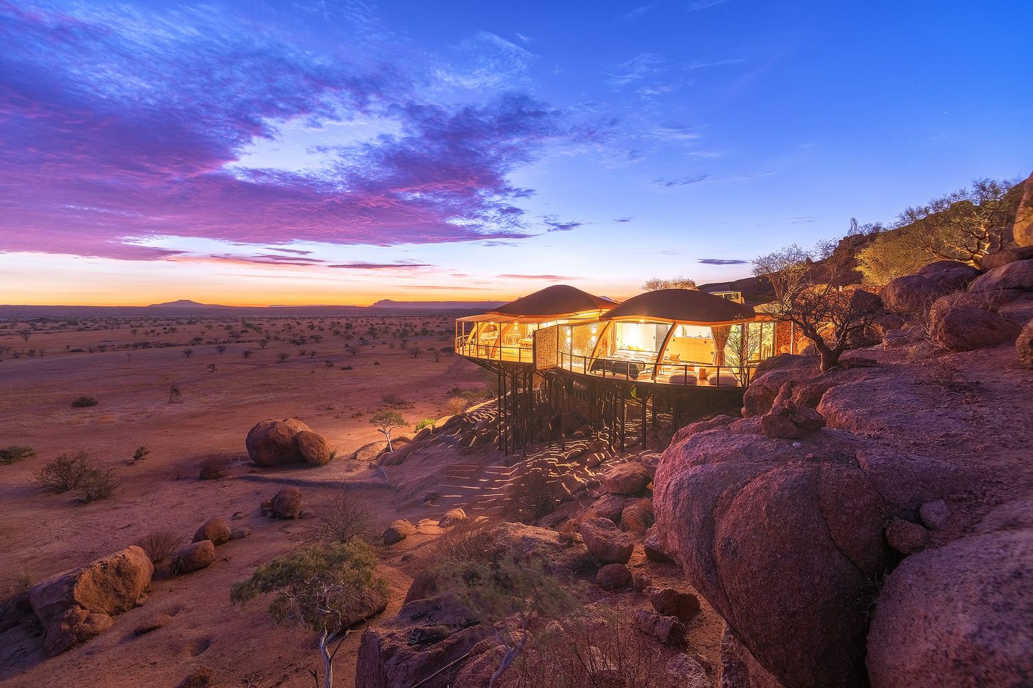 Luxury desert lodge with glass walls nestled among boulders at sunset, overlooking a vast Namibian landscape.