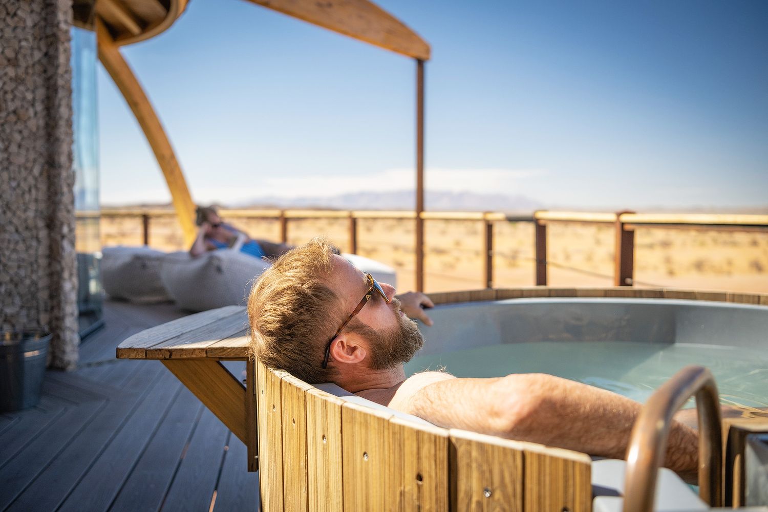 Man relaxing in an outdoor wooden hot tub with a scenic desert landscape view on a sunny day.