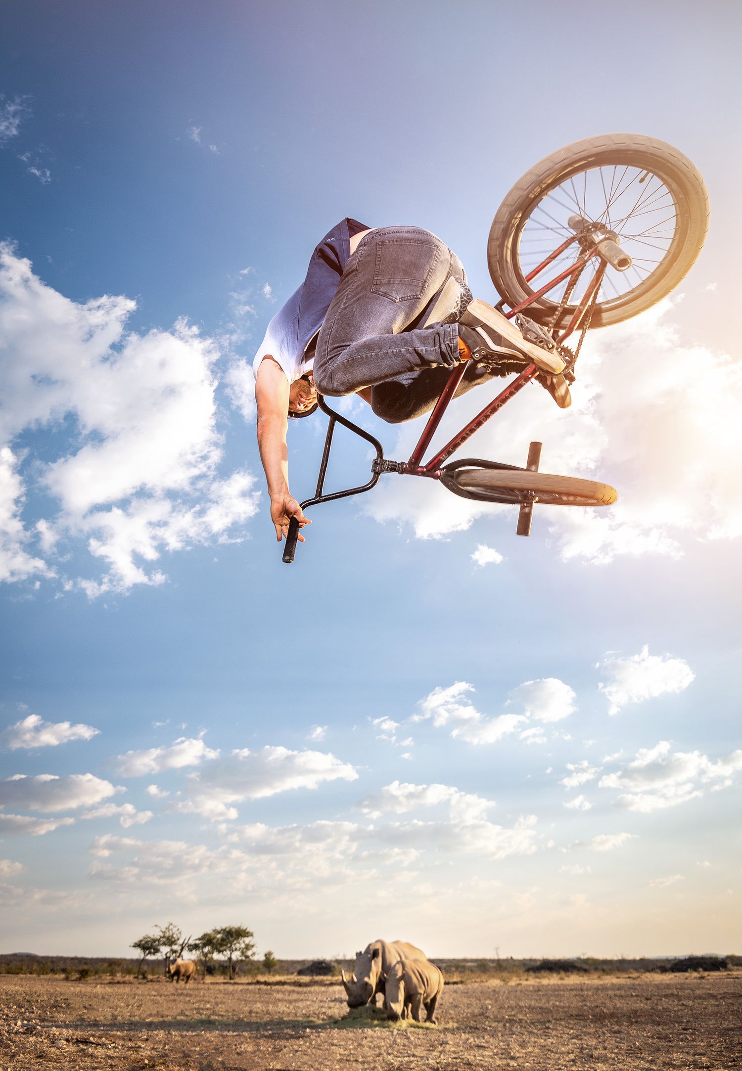 BMX rider performs an aerial trick high above a rhino grazing in a vast African savanna landscape.
