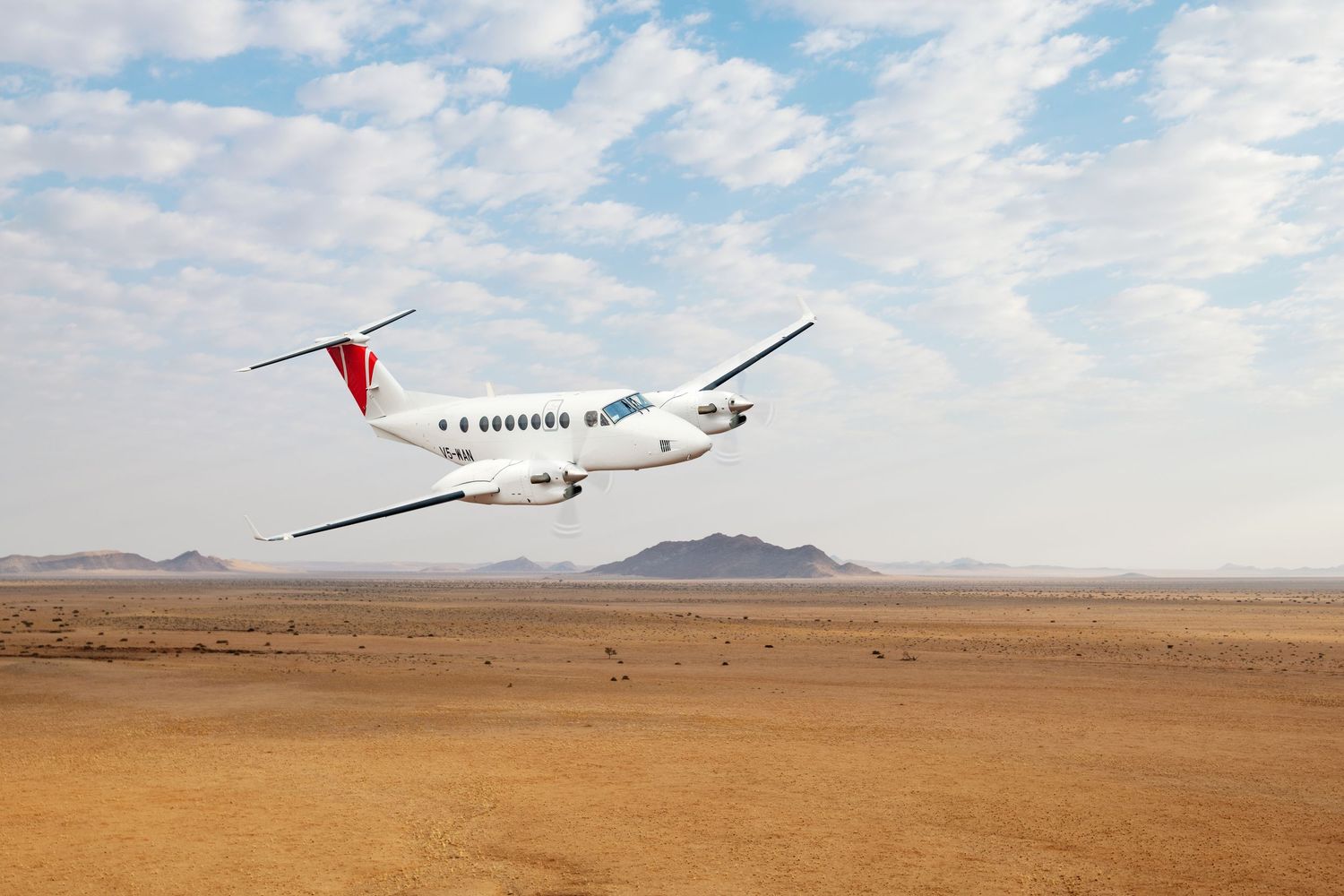 A white regional aircraft with red tail markings flies low over an arid desert landscape under a partly cloudy sky.