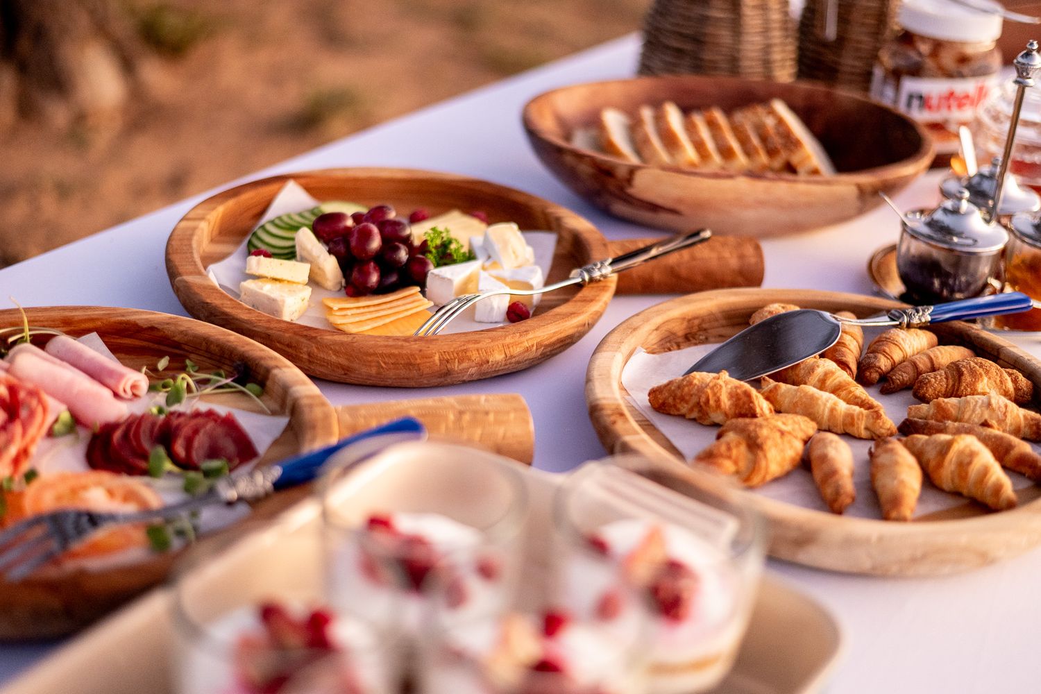 Outdoor safari breakfast spread with croissants, charcuterie, fresh fruit, and Nutella on rustic wooden platters.