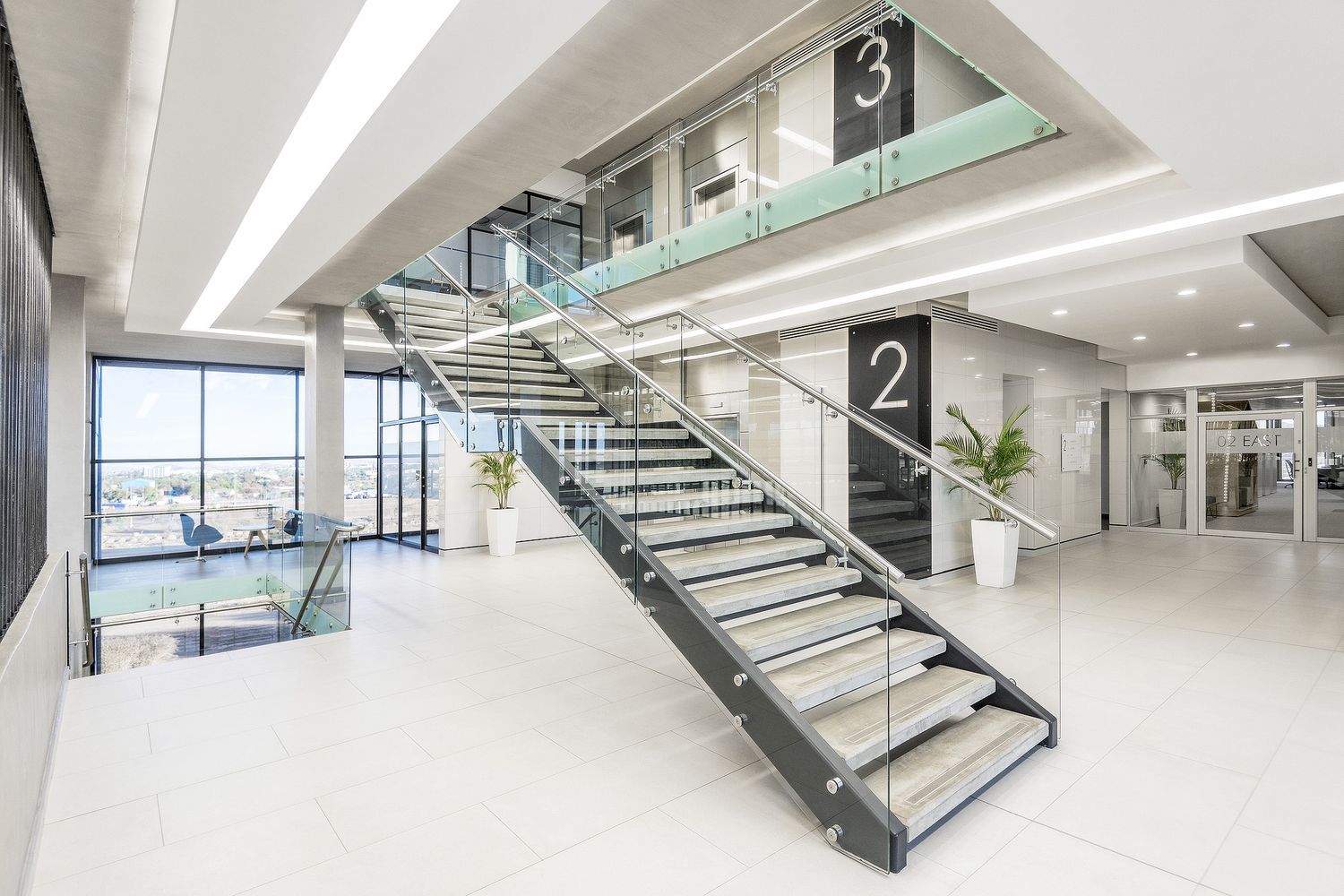 Modern multi-story office interior with open staircase, glass railings, white floors, and large panoramic windows.