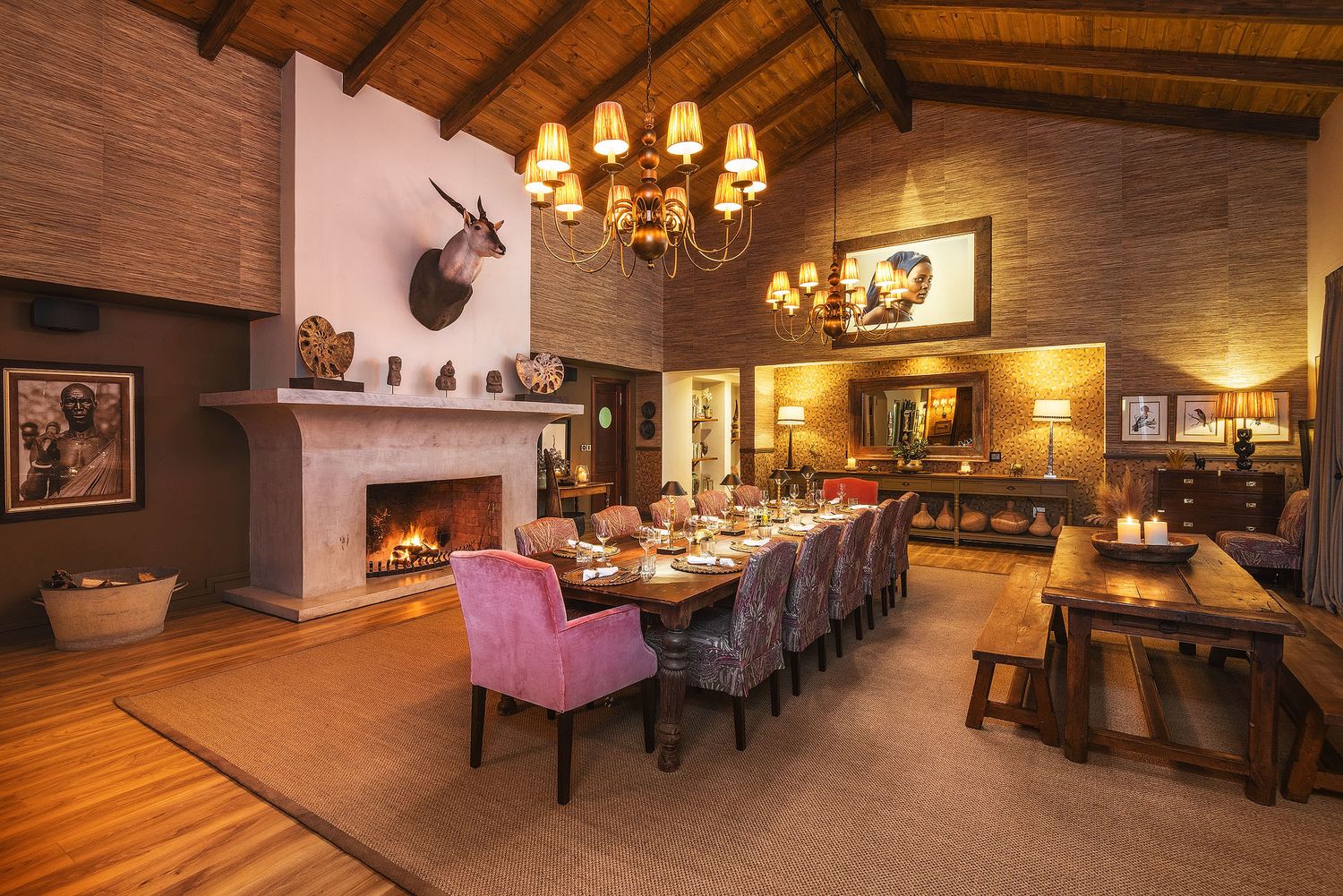 Elegant safari lodge dining room with long table, purple chairs, stone fireplace, chandelier, and rustic wooden decor.