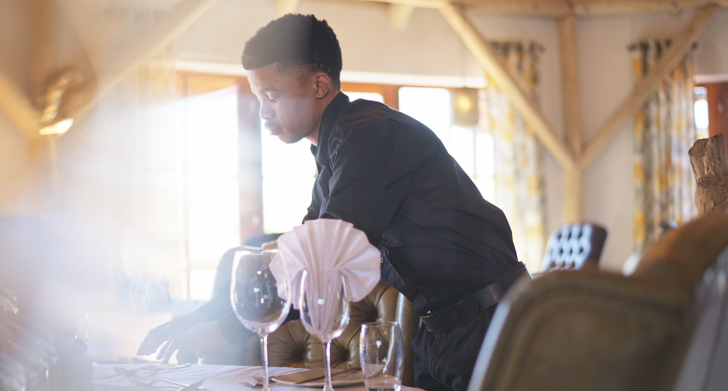 A waiter in formal attire sets an elegant table with wine glasses and decorative napkins in a bright restaurant.