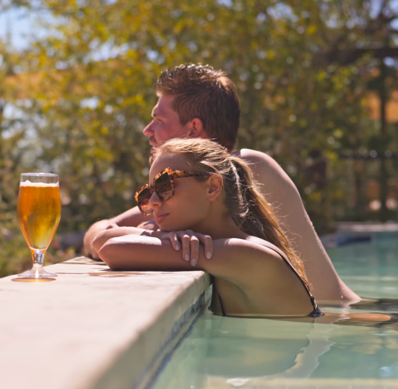 Couple relaxing in a pool leaning on the edge with a glass of beer on a sunny day.