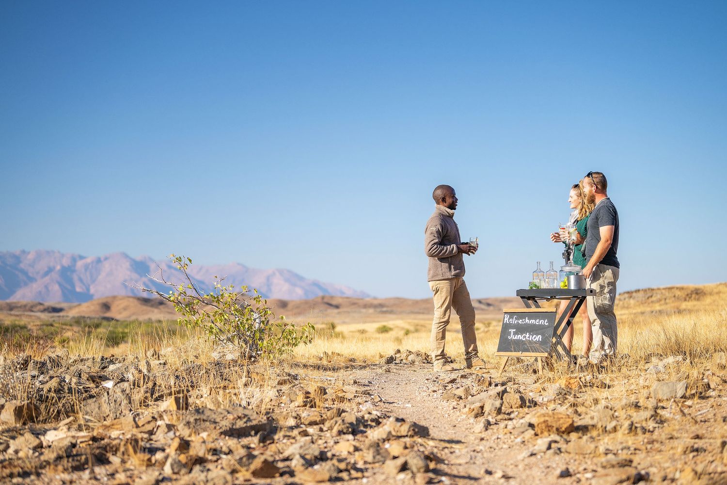 Two people stand on a desert trail with mountain views under a clear blue sky in an arid landscape.