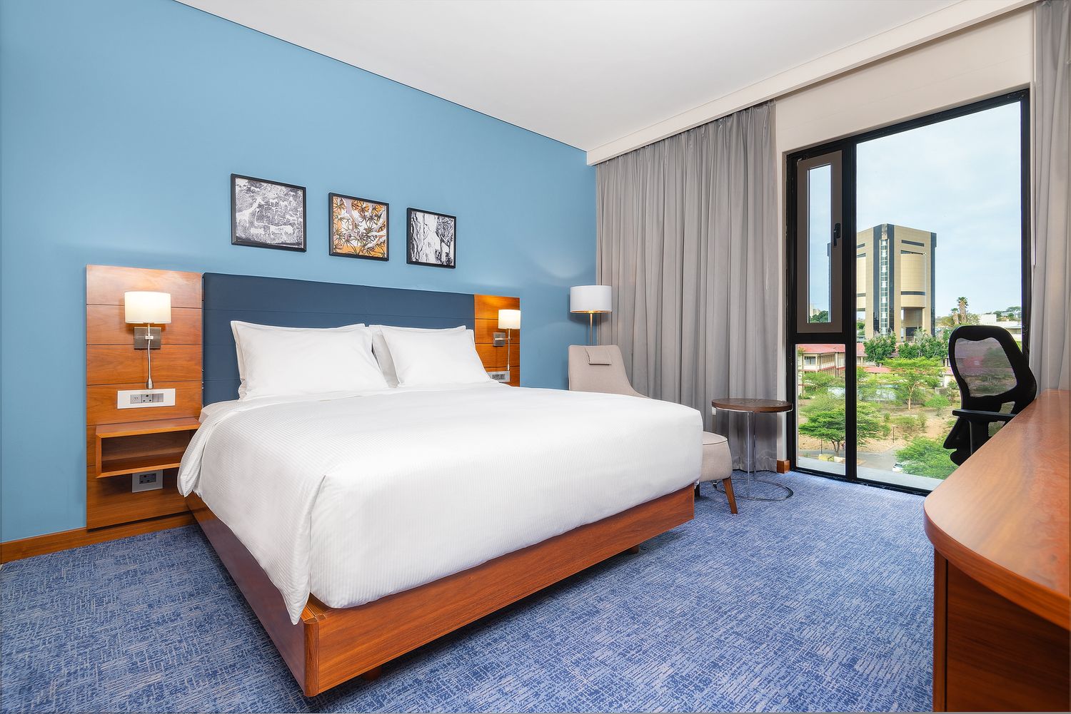 Modern hotel room with king bed, blue accent wall, artwork, large window, and blue carpet flooring.