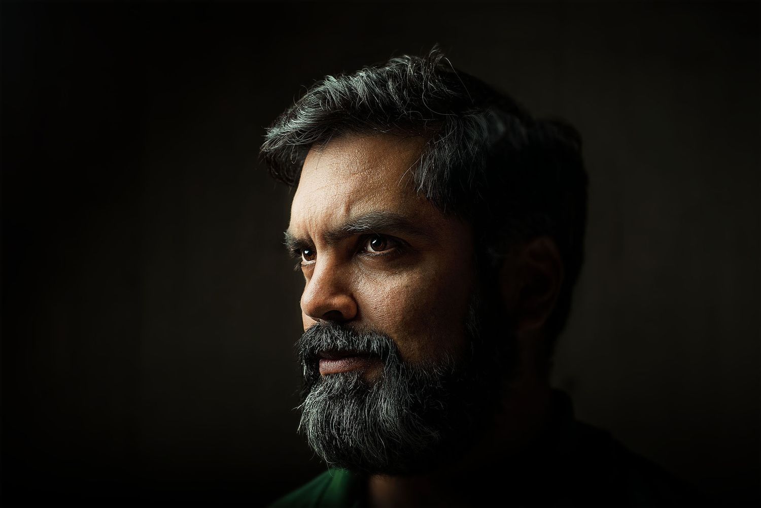 A bearded middle-aged man with dark hair poses in dramatic low-key studio lighting against a dark background.