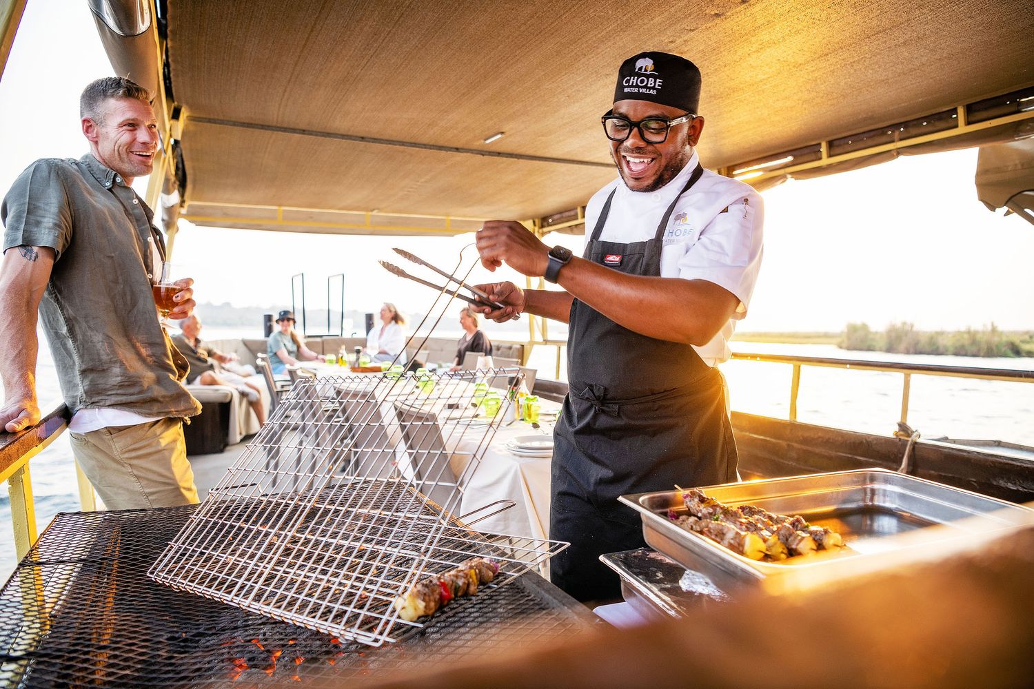 Smiling chef grilling food on outdoor barbecue at sunset on a boat deck with golden light.