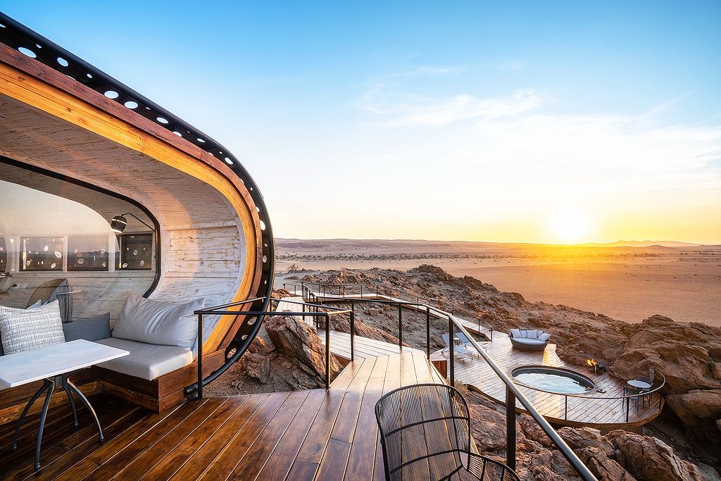 Curved glass luxury suite with open terrace, lounge seating, and breathtaking ocean sunset views over rocky coastline.