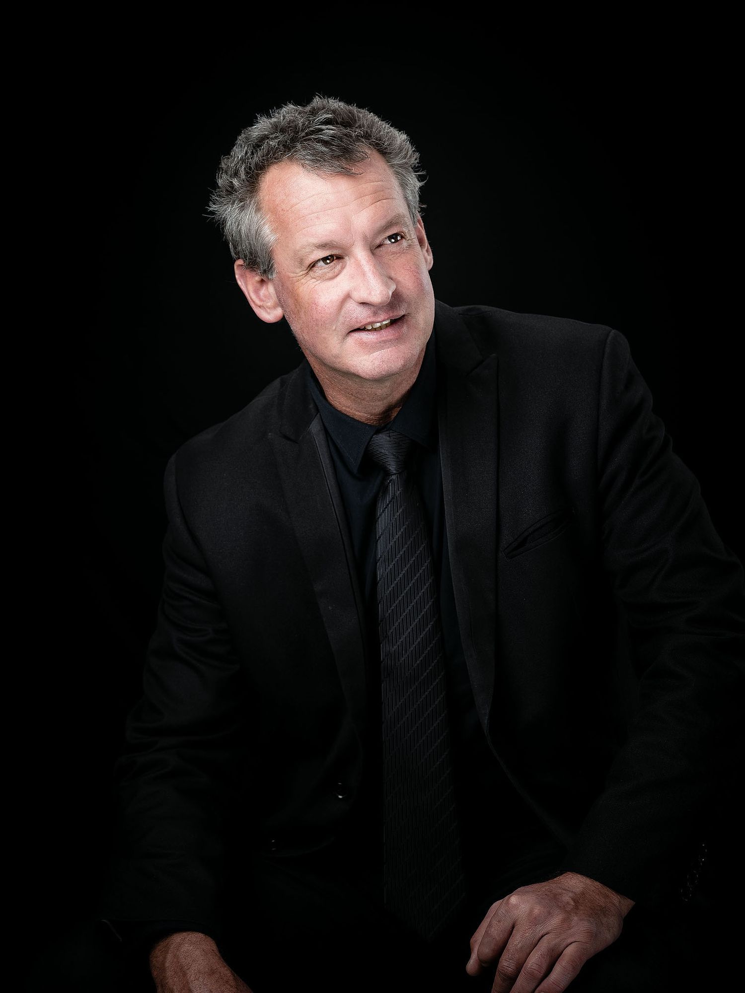 Professional man in black suit and tie posing confidently against dark background in formal portrait photo.