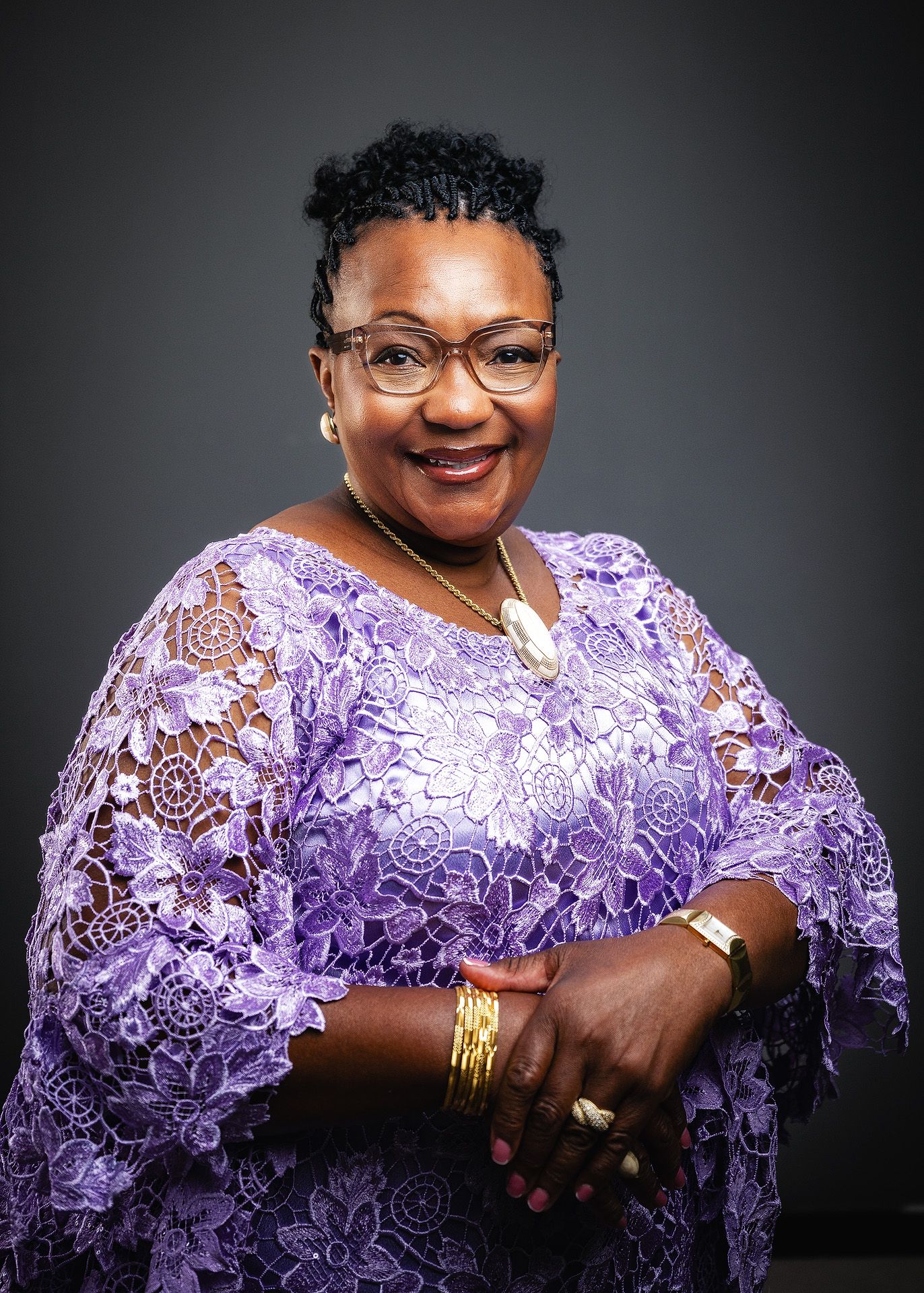 Smiling woman in purple lace dress with glasses and gold jewelry poses confidently for a professional portrait.