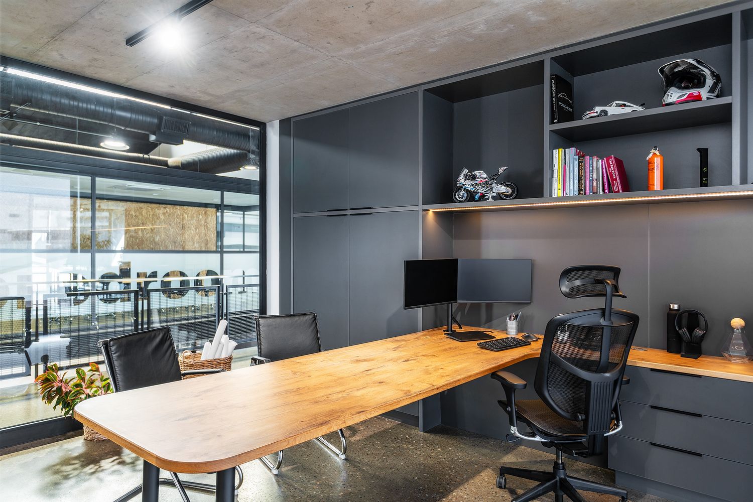 Modern industrial office with wooden desk, ergonomic chair, dark built-in shelving, and glass wall overlooking open workspace.