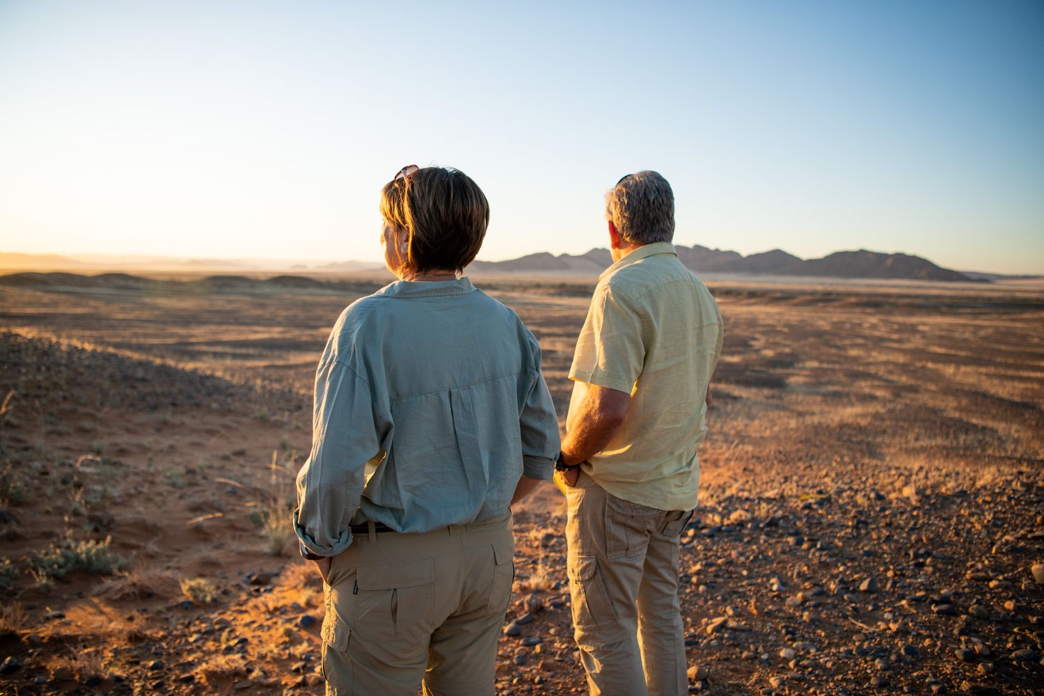 Two people stand back-to-back admiring a vast desert landscape at sunset with distant mountains on the horizon.