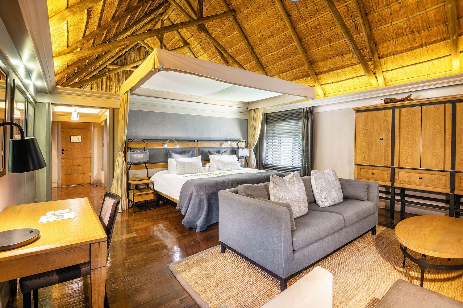 A spacious safari lodge room with thatched ceiling, king bed, sofa, and warm wooden furnishings.