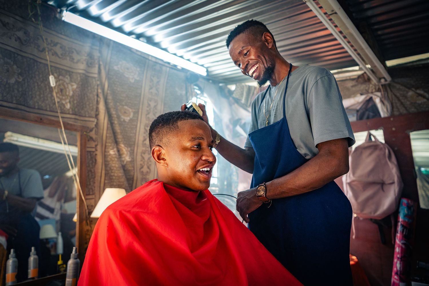 A smiling barber in a blue apron gives a haircut to a young man wearing a red cape in a barbershop.