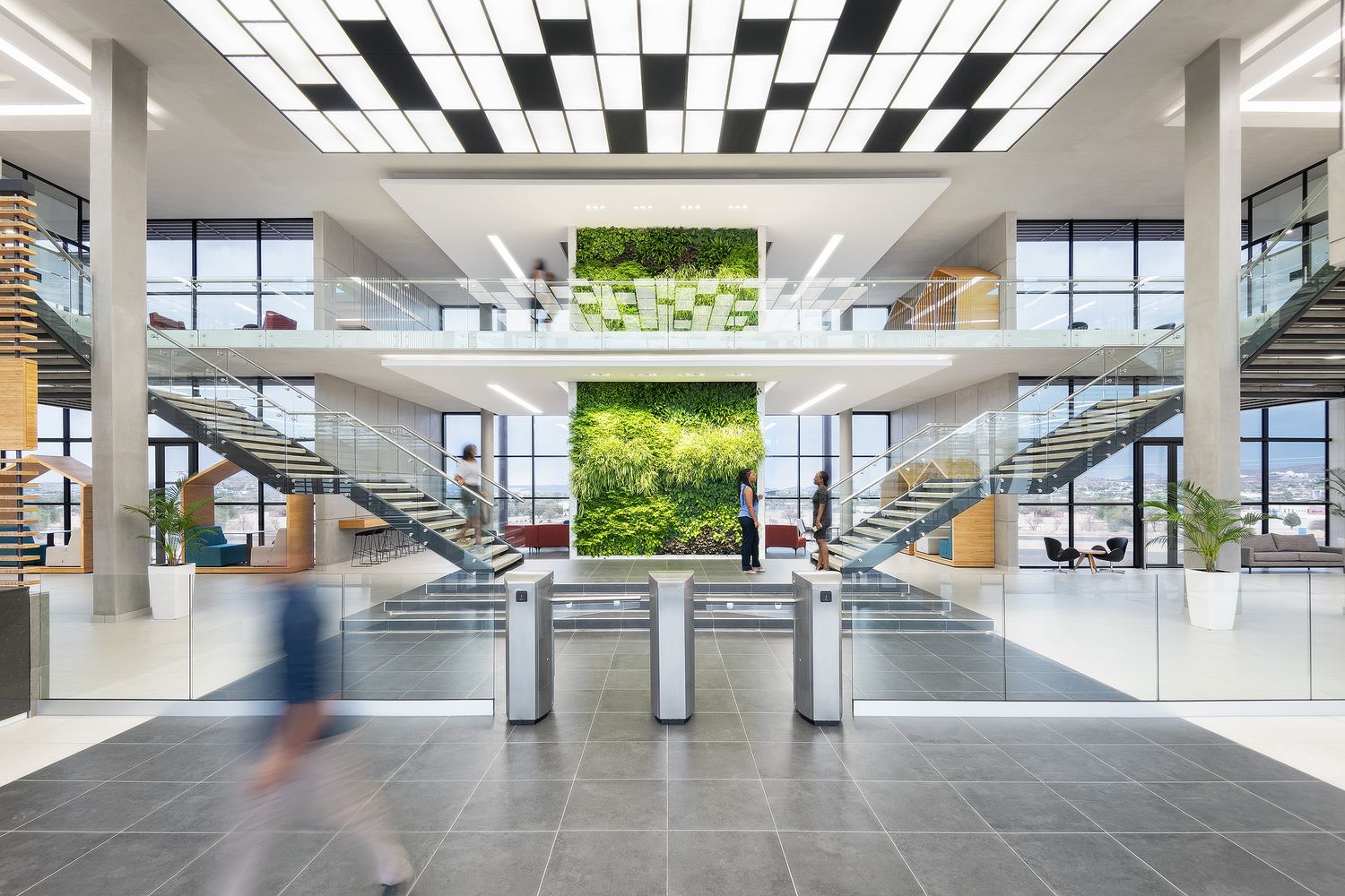 Modern office lobby with green living wall, twin staircases, turnstile gates, and bright grid ceiling lights.