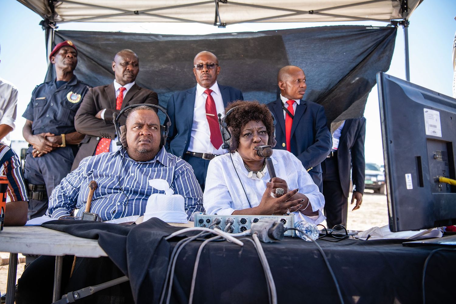 Two people wearing headsets sit at an outdoor audio mixing table during a formal event with officials standing behind.