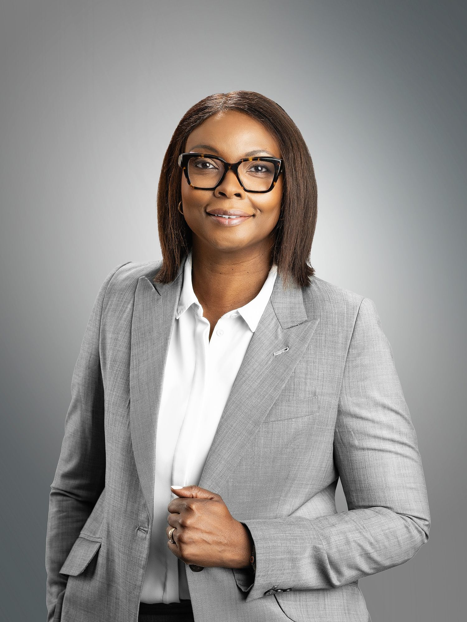 Professional woman in gray blazer and glasses smiling confidently against a neutral studio background.