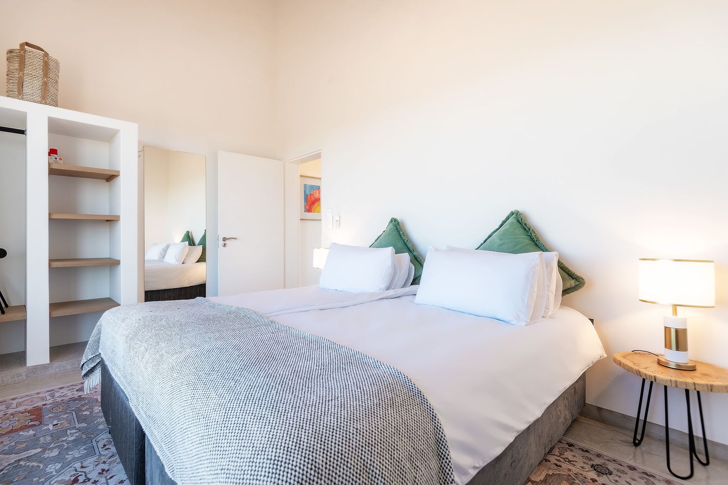 Bright bedroom with a large bed, white bedding, green pillows, bedside lamp, and open shelving near a white door.