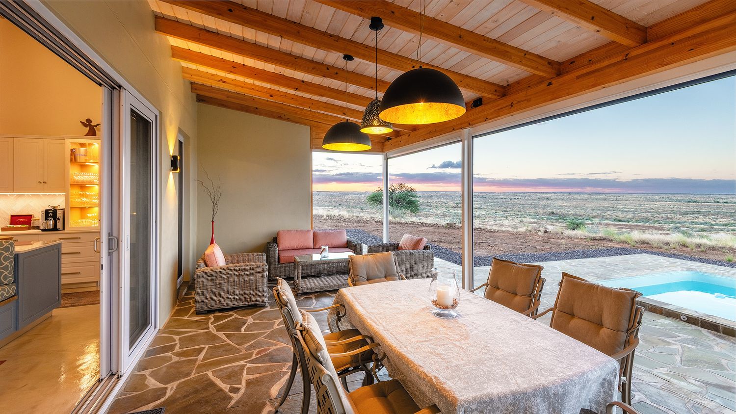 Covered outdoor dining area with pendant lights, wicker furniture, and panoramic sunset views over a pool and landscape.