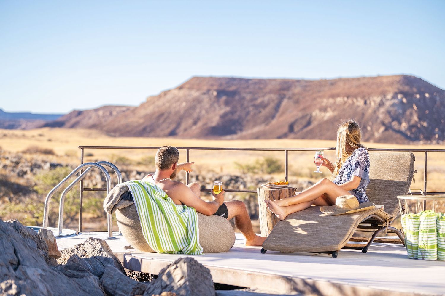 Couple relaxing on lounge chairs by a desert pool with scenic mountain views under a clear blue sky.