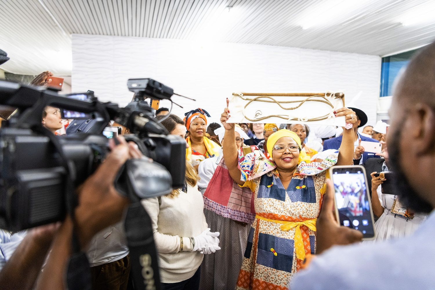 Vibrant African celebration with woman in traditional colorful attire raising hands while being filmed by camera crew.