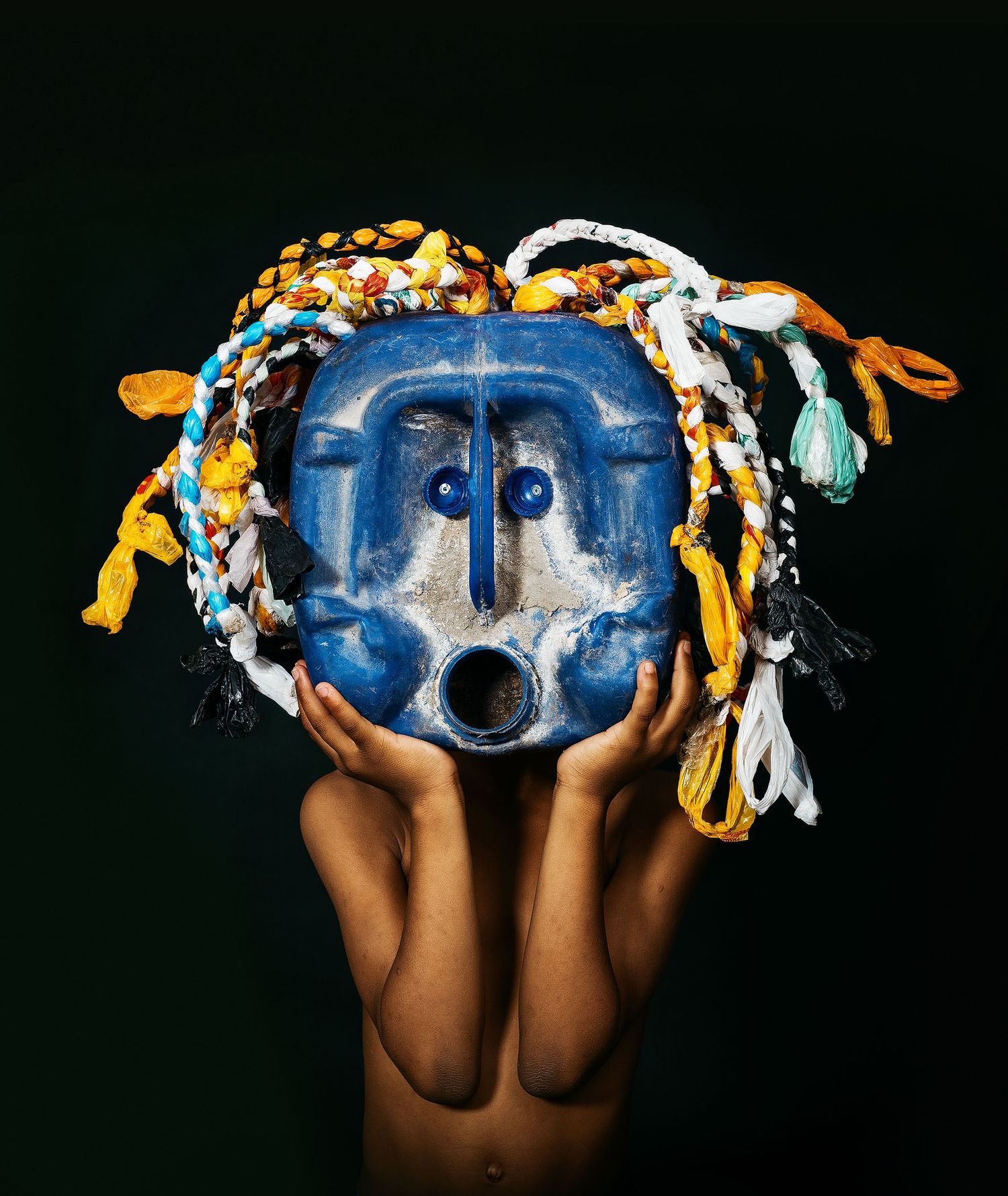 Hands holding a blue ceremonial mask adorned with colorful braided fabric strands against a dark background.