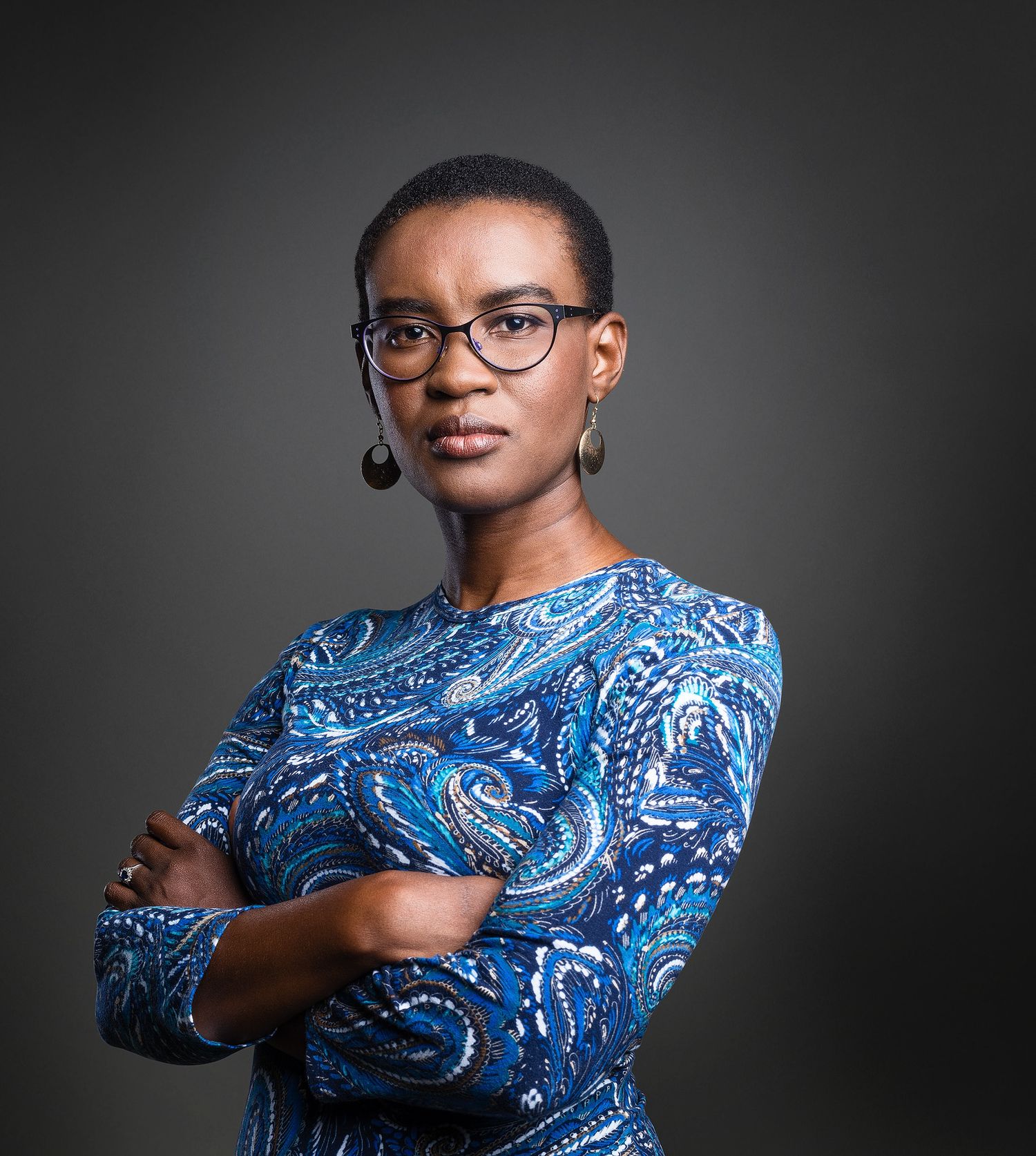 Professional woman in blue paisley dress with arms crossed, wearing glasses, against dark background.