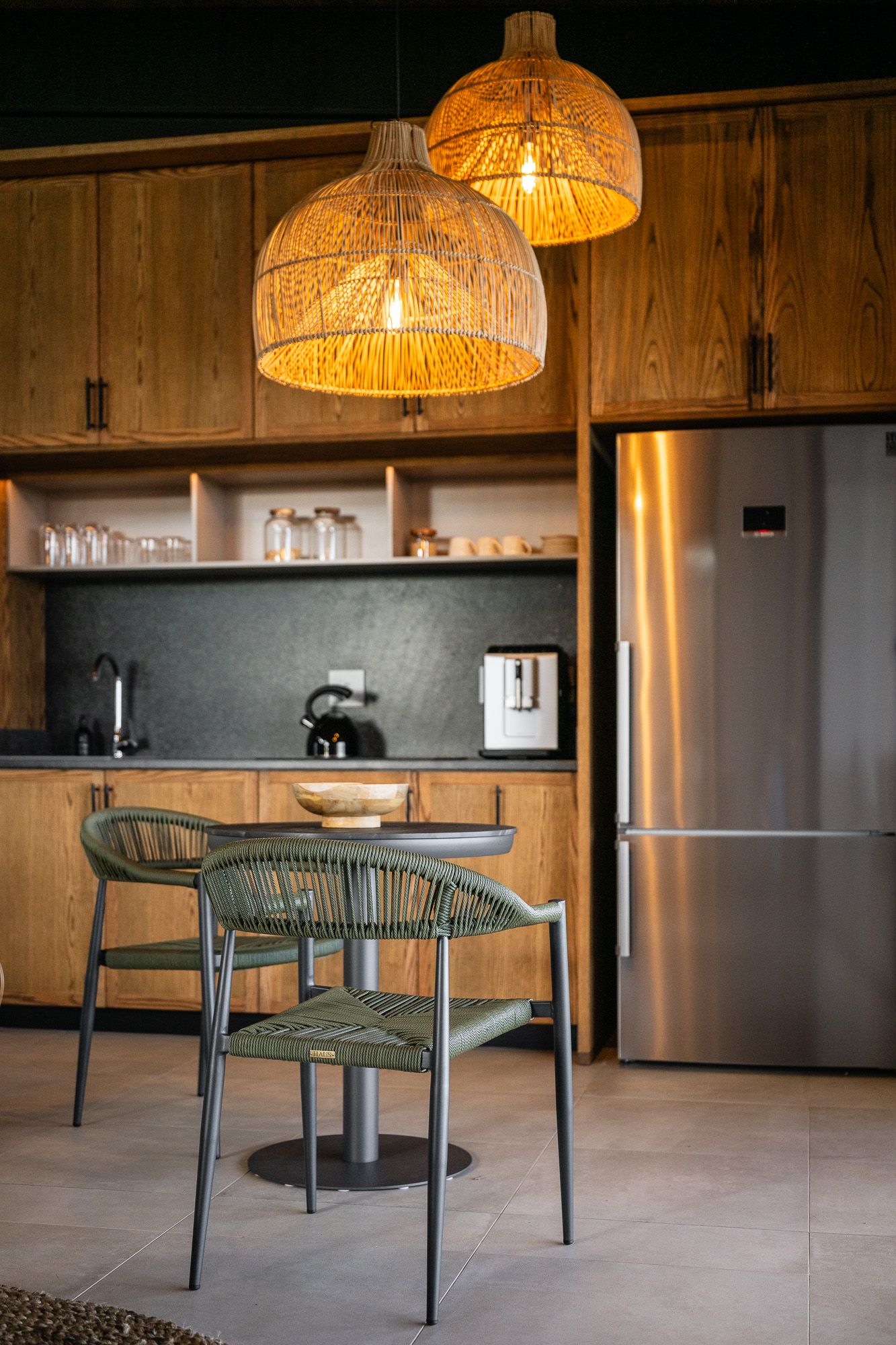Modern kitchen with wicker pendant lights, wooden cabinets, green chairs, and stainless steel refrigerator.