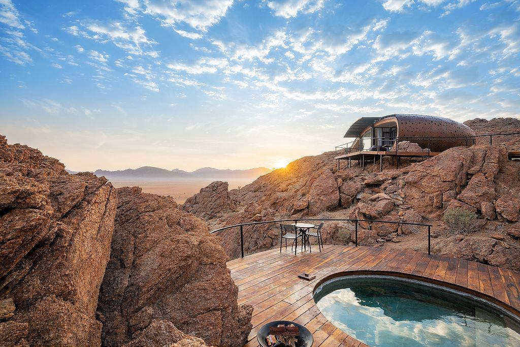 Futuristic curved lodge suite nestled between dramatic desert rocks with infinity pool and panoramic sunset views.