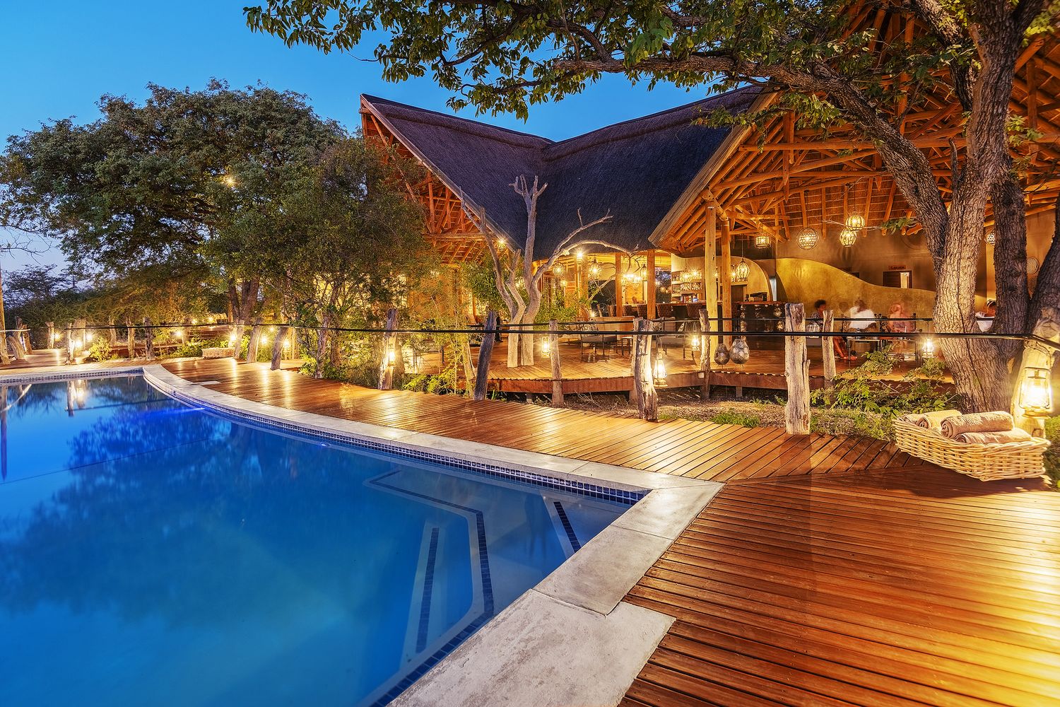 Luxury safari lodge pool deck at dusk with warm lighting, wooden decking, and thatched roof bar area surrounded by trees.