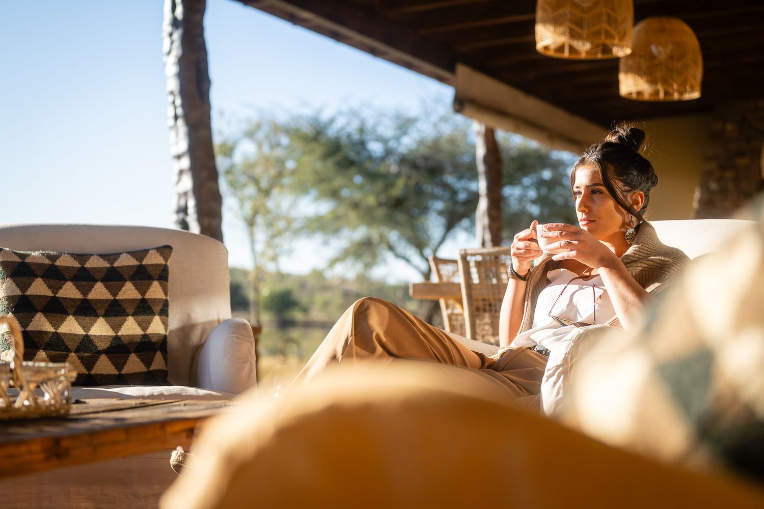 A woman relaxes on a luxury safari lodge lounge, sipping coffee while enjoying warm golden sunlight and scenic outdoor views.