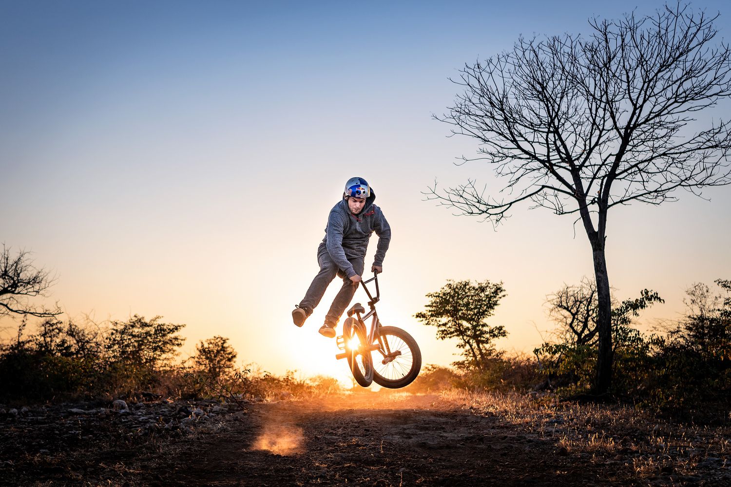 BMX rider performs a dramatic jump silhouetted against a vibrant sunset sky with bare trees in the background.