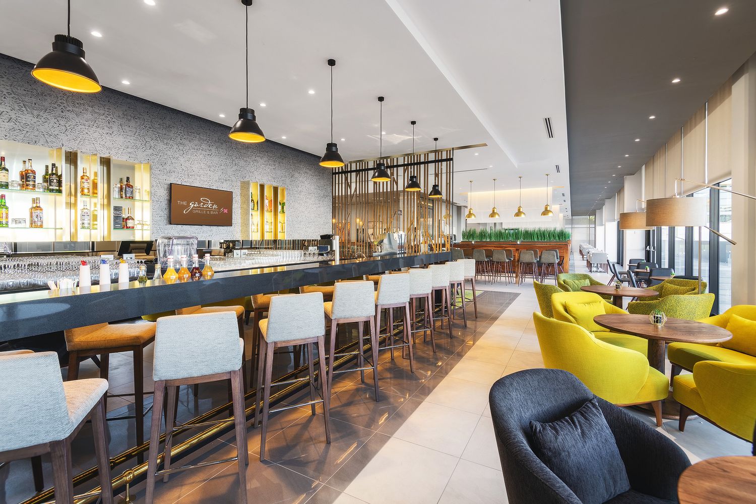Modern hotel bar with white bar stools, yellow accent chairs, pendant lights, and a fully stocked backlit bar in an elegant lounge.